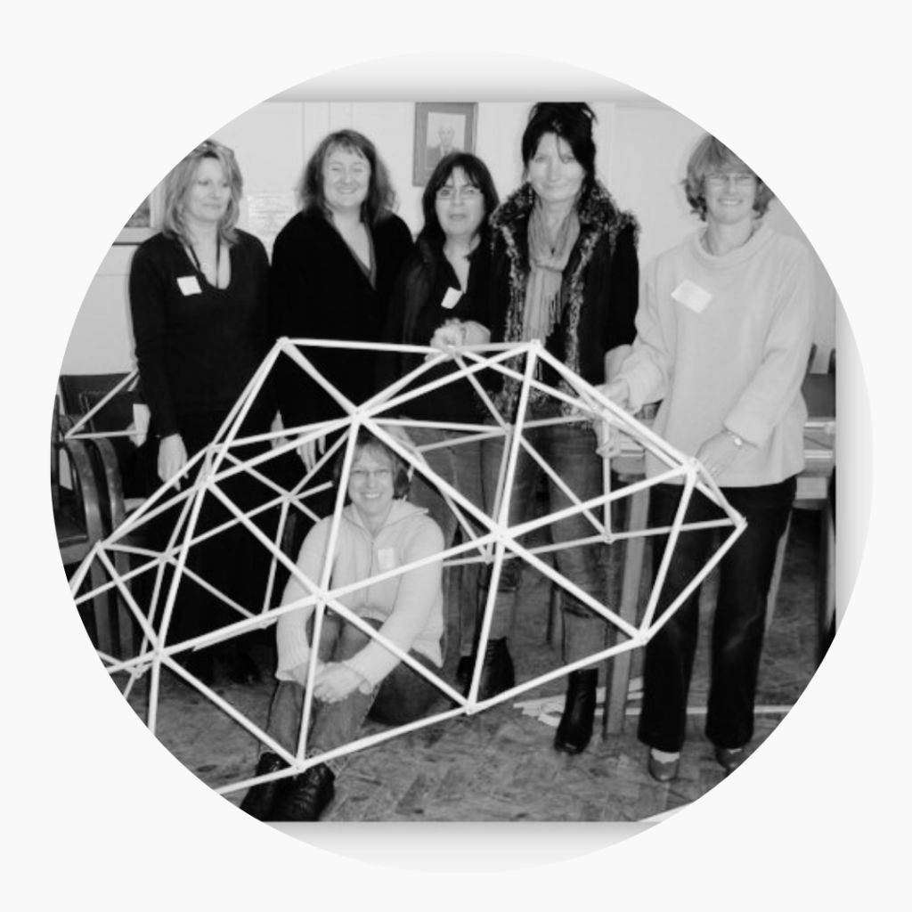 A black and white photo of a group of women standing around and one inside a structure made of rolled and bolted together paper tubes forming a shelter